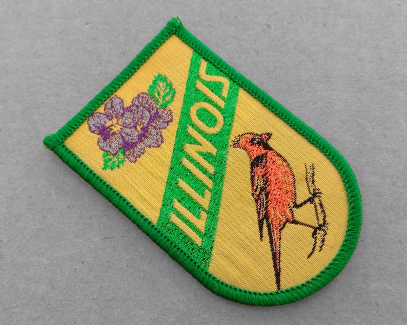 May include: A yellow and green embroidered patch with the word "ILLINOIS" in green letters and a purple flower and a red bird on a branch.