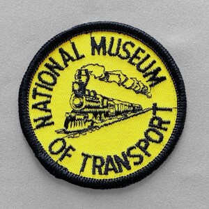 May include: Round yellow patch with black embroidered text and border. The patch reads "NATIONAL MUSEUM OF TRANSPORT" around the edge. A black train is in the center, with smoke billowing from the smokestack.