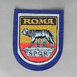May include: A blue patch with a white border featuring a shield design. The shield displays the word "ROMA" above an image of a wolf nursing two babies, with the letters "SPQR" below. The background is gold and red.