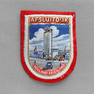 May include: A rectangular patch with a red border and a detailed image of a tall structure, sailboats, and the text "AFSLUITDIJK HOLLAND-FRIESLAND". The image is set against a blue sky with white clouds.