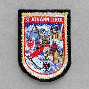 May include: Embroidered patch with a black border and gold stitching. The patch features a skier, buildings, and the text "ST. JOHANN TIROL" in red. The patch also includes the text "TIROL" and "AUSTRIA".