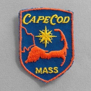 May include: Embroidered patch featuring a shield design with the words "CAPE COD MASS" in yellow. The patch has a blue background with an orange outline and an orange Cape Cod outline and starburst.