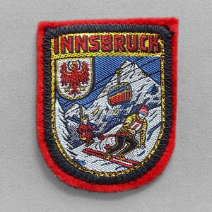 May include: Embroidered patch with the word "INNSBRUCK" at the top. The patch features a skier, a cable car, and a mountain scene with a red border and black trim.