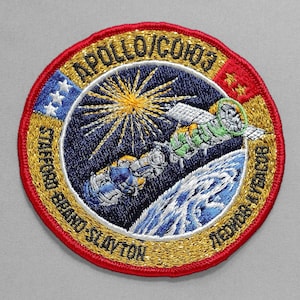 May include: Embroidered patch featuring the Apollo-Soyuz Test Project mission. The circular patch has a red border with gold trim. The central design depicts a spacecraft, Earth, and a sunburst against a dark blue background. The names of the astronauts are also included.