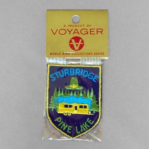 May include: A vintage patch from the Voyager World Wide Collectors Series. The patch features a blue shield design with the words "Sturbridge" and "Pine Lake" in yellow and blue. The patch has a yellow border and a design of a yellow bus, trees, and a green hat.