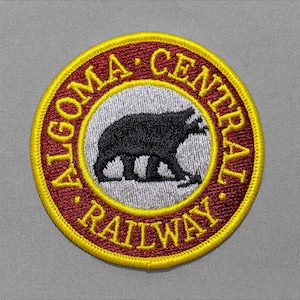 May include: A circular embroidered patch featuring a black bear silhouette centered on a light gray background. A deep red border surrounds the center, containing the yellow text ALGOMA CENTRAL RAILWAY. The entire patch has a bright yellow edge.