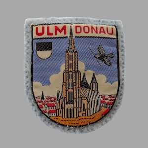 May include: A light blue patch with a detailed illustration of a cathedral, a flying bird, and the text "ULM DONAU" in red. The patch has a light blue, textured border and a small flag icon.