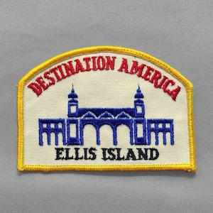 May include: Embroidered patch featuring the words "DESTINATION AMERICA" in red, arched across the top. Below, a blue illustration of Ellis Island is centered, with the words "ELLIS ISLAND" in black at the bottom. The patch has a yellow border.