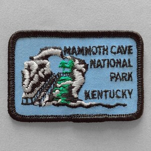 May include: Embroidered patch featuring the words "Mammoth Cave National Park Kentucky" in black text on a light blue background. The patch has a brown border and a detailed illustration of the cave entrance in white, black, and green.
