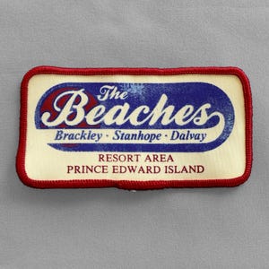 May include: A rectangular patch with a cream-colored background and a red border. The patch features the text "The Beaches" in a blue and red design, with the words "Brackley - Stanhope - Dalvay" below. The text "Resort Area Prince Edward Island" is also included.