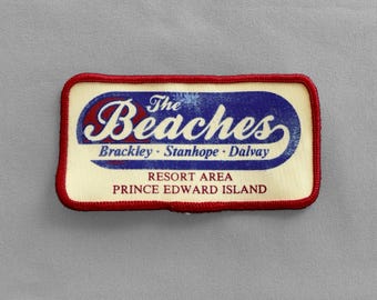 May include: A rectangular patch with a cream-colored background and a red border. The patch features the text "The Beaches" in a blue and red design, with the words "Brackley - Stanhope - Dalvay" below. The text "Resort Area Prince Edward Island" is also included.