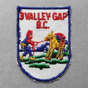 May include: Embroidered patch with a shield shape, featuring the text "3 VALLEY GAP B.C." in red. The design includes a figure in blue and red pulling a yellow animal with a red head covering. Green and blue landscape elements are also present.