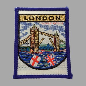 Puede incluir: Parche rectangular con borde azul, que representa el Tower Bridge de Londres. El parche incluye el texto "LONDON TOWER BRIDGE" y las banderas de Inglaterra y el Reino Unido. El diseño es tejido.