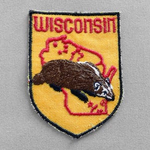 May include: A yellow and red embroidered patch with the word "WISCONSIN" in red lettering. The patch features a brown badger inside the outline of the state of Wisconsin.