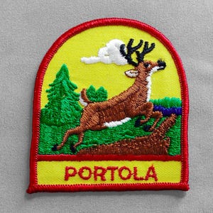 May include: Embroidered patch featuring a brown deer leaping over a log, with a green tree and cloud on a yellow background. The patch has a red border and the word "PORTOLA" in red letters.