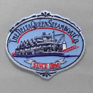 May include: Embroidered patch featuring a steamboat design with the text "THE DELTA QUEEN STEAMBOAT CO." and "SINCE 1890." The patch has a light blue background with a white and navy blue border. The steamboat is white and navy blue.