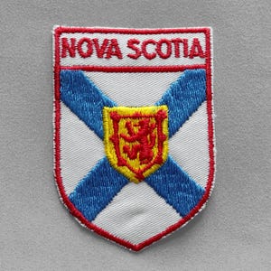 May include: Embroidered patch featuring the Nova Scotia provincial flag. The patch has a white background with a blue saltire, a red lion on a yellow shield, and the words "NOVA SCOTIA" in red.