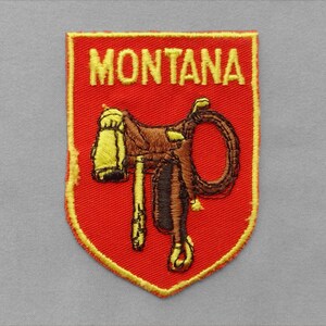 May include: A shield-shaped embroidered patch with a red background and a yellow border. The word MONTANA is stitched in yellow at the top. Centered below is a brown western saddle with yellow accents and a coiled rope attached to the side.