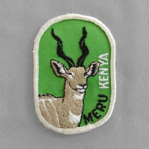 May include: Green embroidered patch with a white outline featuring a brown and white gerenuk with long black horns. The text "MERU KENYA" is embroidered in white on the patch.