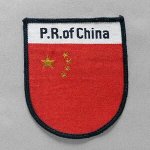 May include: A shield-shaped embroidered patch featuring a red background with five gold stars arranged in an arc. The top section is white with the black text P.R. of China printed across it. The entire patch is bordered by a thick black stitched edge.