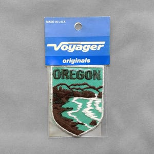 May include: A shield-shaped embroidered patch featuring a landscape of Oregon with brown mountains, a teal sky, and white water. The patch is inside a clear plastic sleeve with a blue header card that reads Voyager originals and MADE IN U.S.A.
