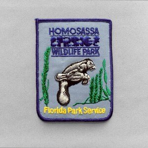 May include: A rectangular embroidered patch with a light blue background and a dark blue border. It features a grey manatee illustration, green seaweed, and the text HOMOSASSA SPRINGS WILDLIFE PARK in blue and Florida Park Service in yellow.