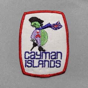 May include: A rectangular embroidered patch with a red border on a white background. It features a green turtle wearing a black hat and a purple jacket, holding a blue and red flag. The text "cayman ISLANDS" is embroidered in blue below.