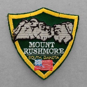 May include: Embroidered patch featuring Mount Rushmore in white against a green background. The patch has a yellow border and includes the text "MOUNT RUSHMORE SOUTH DAKOTA" and a small American flag.