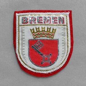 May include: Embroidered patch with the word "BREMEN" at the top. The patch features a red shield with a silver key and a gold crown. The patch is bordered with red and gold trim.