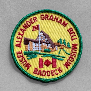 May include: Embroidered patch with a yellow background and green border. The patch features the text "ALEXANDER GRAHAM BELL MUSEUM BADDECK" in a circular pattern. A detailed illustration of a building, trees, and a Canadian flag is in the center.