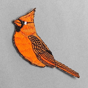May include: Embroidered patch of a cardinal bird in shades of orange and black. The bird is depicted in profile, with detailed stitching creating a textured appearance. The patch is set against a gray background.