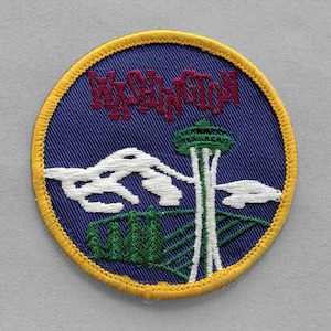 May include: Embroidered patch featuring the word "WASHINGTON" in red, above a depiction of the Space Needle, mountains, and trees. The patch has a blue background with a yellow border.