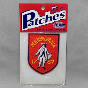 May include: Iron-on patch featuring a red shield with the words "Pennsylvania 1787" in yellow lettering. A white figure of a man in a colonial hat is depicted inside the shield.