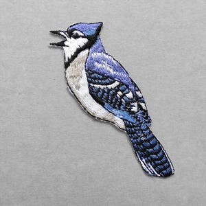 May include: An embroidered patch of a blue jay bird with its beak open. The bird features vibrant blue, white, and black thread detailing, showcasing the characteristic crest, patterned wings, and long tail feathers of the species in a side profile.
