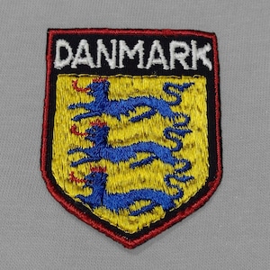 May include: A red, black, and yellow embroidered patch with the word "DANMARK" in white lettering. The patch features three blue lions with red crowns on a yellow background.