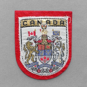 May include: A shield-shaped embroidered patch with a red felt border. The center features the Canadian coat of arms with a lion and unicorn, topped by the word CANADA in black letters. It includes a small Canadian flag and a blue flag.