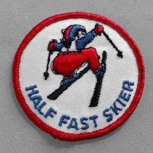 May include: A white embroidered patch with a red border. The patch features a cartoon skier in a red and blue suit, skiing down a slope. The text "Half Fast Skier" is written in blue around the skier.