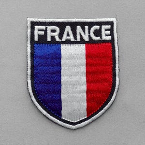 May include: A shield-shaped embroidered patch featuring the word FRANCE in white capital letters on a black background above a vertical tricolor flag design in blue, white, and red. The entire patch is outlined with a thick white stitched border.
