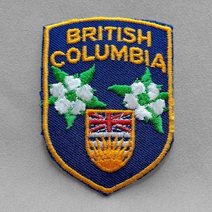 May include: Embroidered patch featuring the words "BRITISH COLUMBIA" in gold lettering on a blue shield. The patch includes floral and crest designs with green, white, red, and yellow accents. The patch has a gold border.