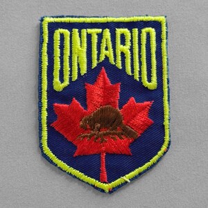 May include: Embroidered patch featuring the word "ONTARIO" in yellow, arched over a red maple leaf with a brown beaver. The patch has a blue background with a yellow border. The patch is rectangular with rounded corners.