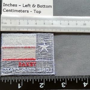 May include: A white, red, and blue embroidered patch with a white star in the center. The patch reads "SAINT" in red letters and "SHOOTERS" in blue letters. The patch is approximately 2 inches wide and 1.5 inches tall.