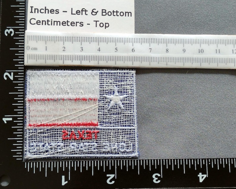 May include: A white, red, and blue embroidered patch with a white star in the center. The patch reads "SAINT" in red letters and "SHOOTERS" in blue letters. The patch is approximately 2 inches wide and 1.5 inches tall.