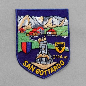 May include: Embroidered patch with a shield design featuring a mountain landscape, buildings, and a stone tower. The patch includes the text "SAN GOTTARDO" and "2114 m" in yellow lettering on a navy blue background.