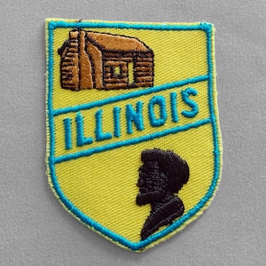 May include: A yellow embroidered patch with a shield shape. The patch features a brown log cabin, the word "ILLINOIS" in teal, and a black silhouette of a person's profile. The patch has a teal border.