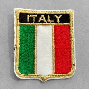May include: Embroidered patch in the shape of a shield, featuring the Italian flag's green, white, and red vertical stripes. The word "ITALY" is embroidered in gold thread on a black background at the top. The patch has a gold border.