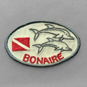 May include: An oval-shaped embroidered patch with an off-white background. It features two dolphins in dark thread, a red and white diver down flag, and the word BONAIRE stitched in red capital letters, all enclosed by a dark border.