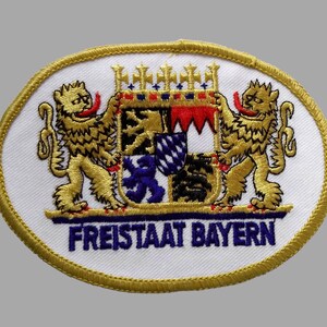 May include: Embroidered patch featuring the Bavarian coat of arms. The design includes two golden lions flanking a shield with a crown, and the text "FREISTAAT BAYERN" in blue. The patch has a gold border and a white background.