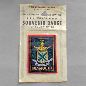 May include: A red, white, and blue woven souvenir badge with the text "GLENTHORNE" SERIES MADE IN ENGLAND, SELF ADHESIVE OR SEW ON, ★★★ WOVEN ★★★, SOUVENIR BADGE OF YOUR VISIT TO PLYMOUTH. The badge features a blue and gold crest with a crown and the text "PLYMOUTH".
