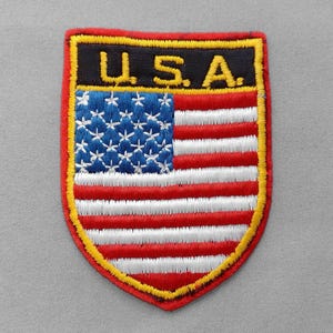 May include: Embroidered patch featuring the American flag design. The patch has a shield shape with a red border and a yellow outline. The top section reads "U.S.A." in yellow letters on a black background. The flag design includes blue with white stars and red and white stripes.
