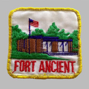 May include: Embroidered patch featuring a building with a blue roof, brown walls, and white windows, set against green trees and a waving American flag. The text "FORT ANCIENT" is in red letters.